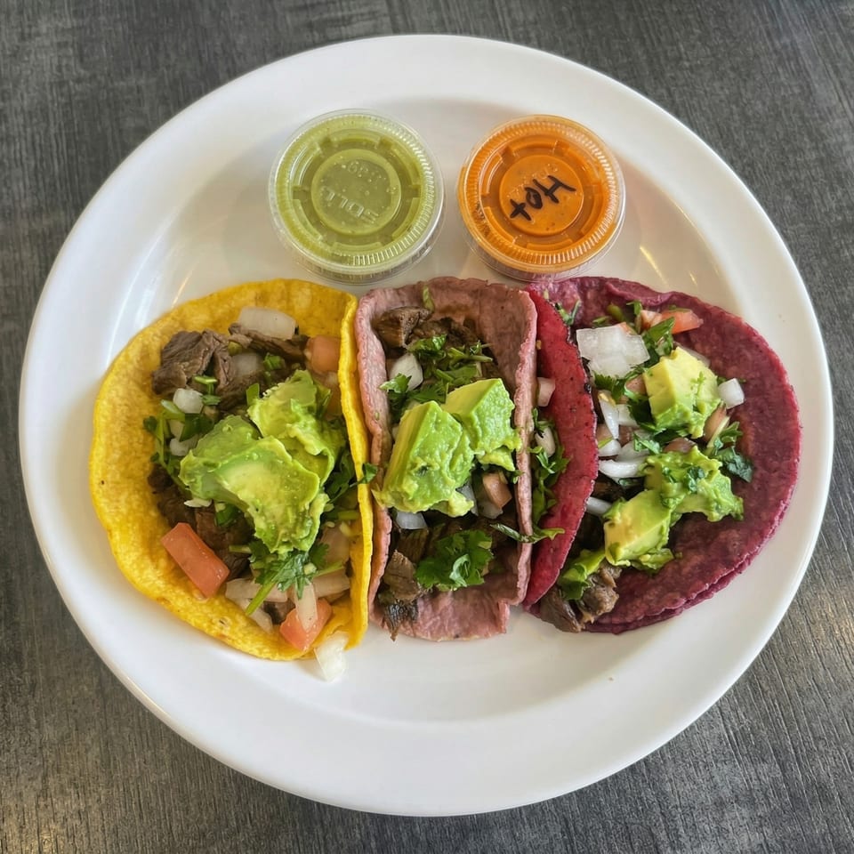 Three colorful tacos with meat, avocado, onions, and cilantro, with two sauce containers on a white plate.