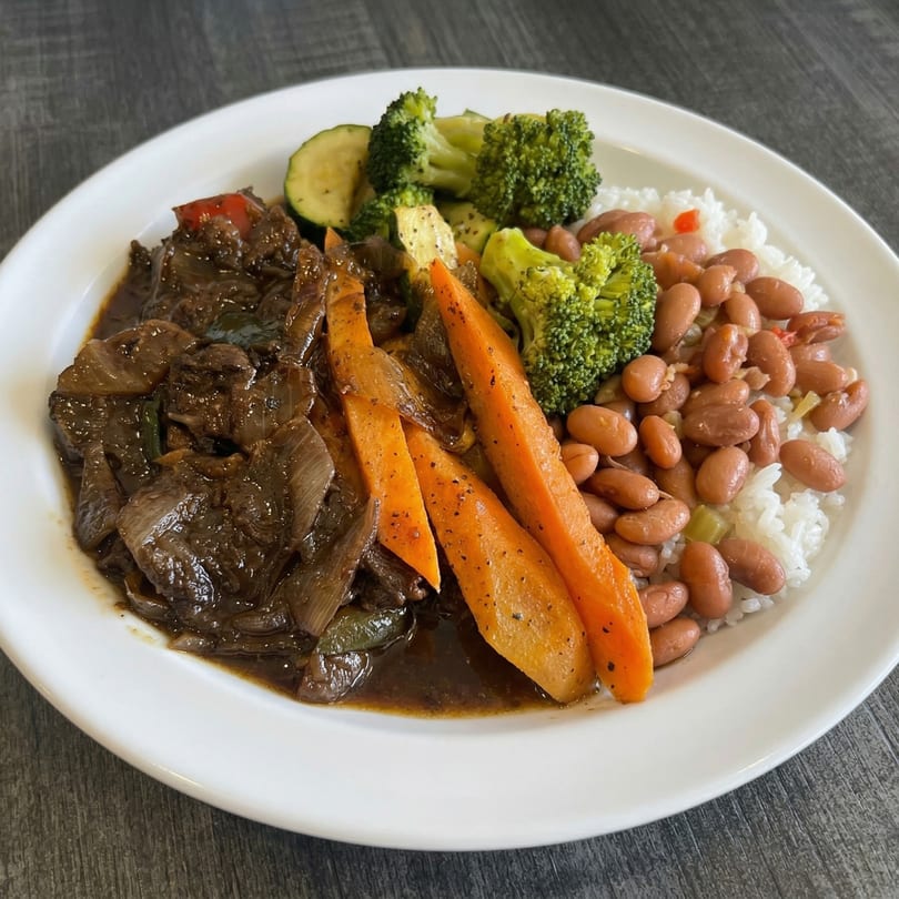 A plate of dark stew with onions, carrots, broccoli, zucchini, white rice, and pinto beans.